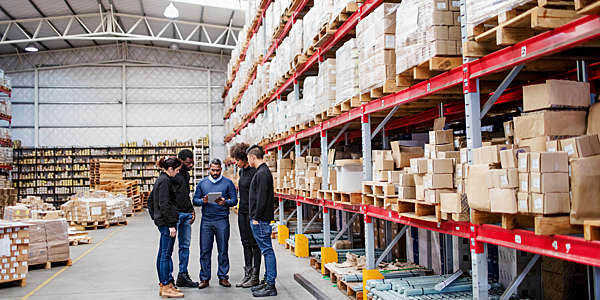 Fiver warehouse workers, one with clipboard, talk shop amidst the storage racks.